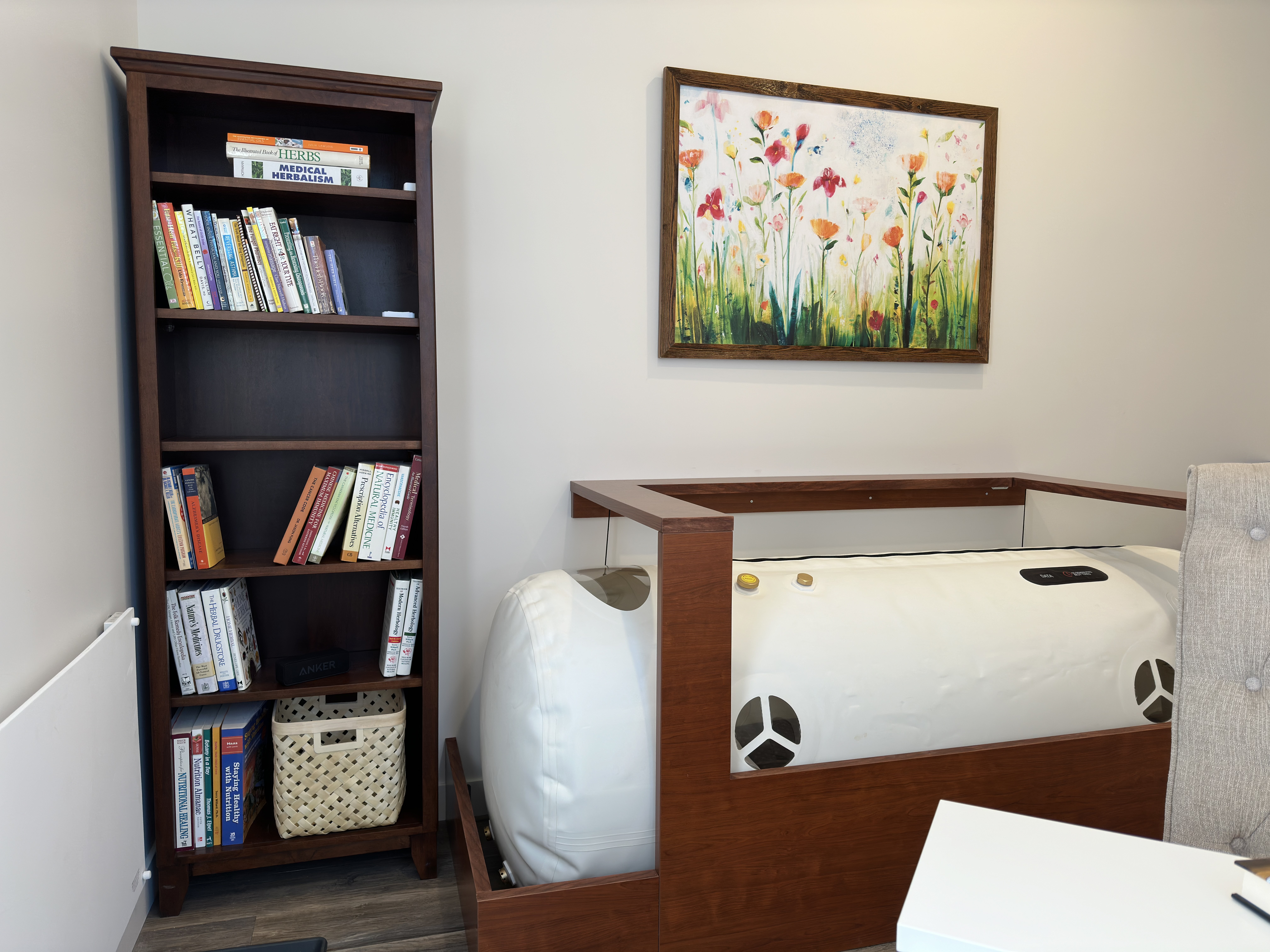 A warm wood-framed soft-shell white hyperbaric chamber in a sunlit room. A tufted upholstered chair sits beside it; a floral painting hangs on the wall above.
