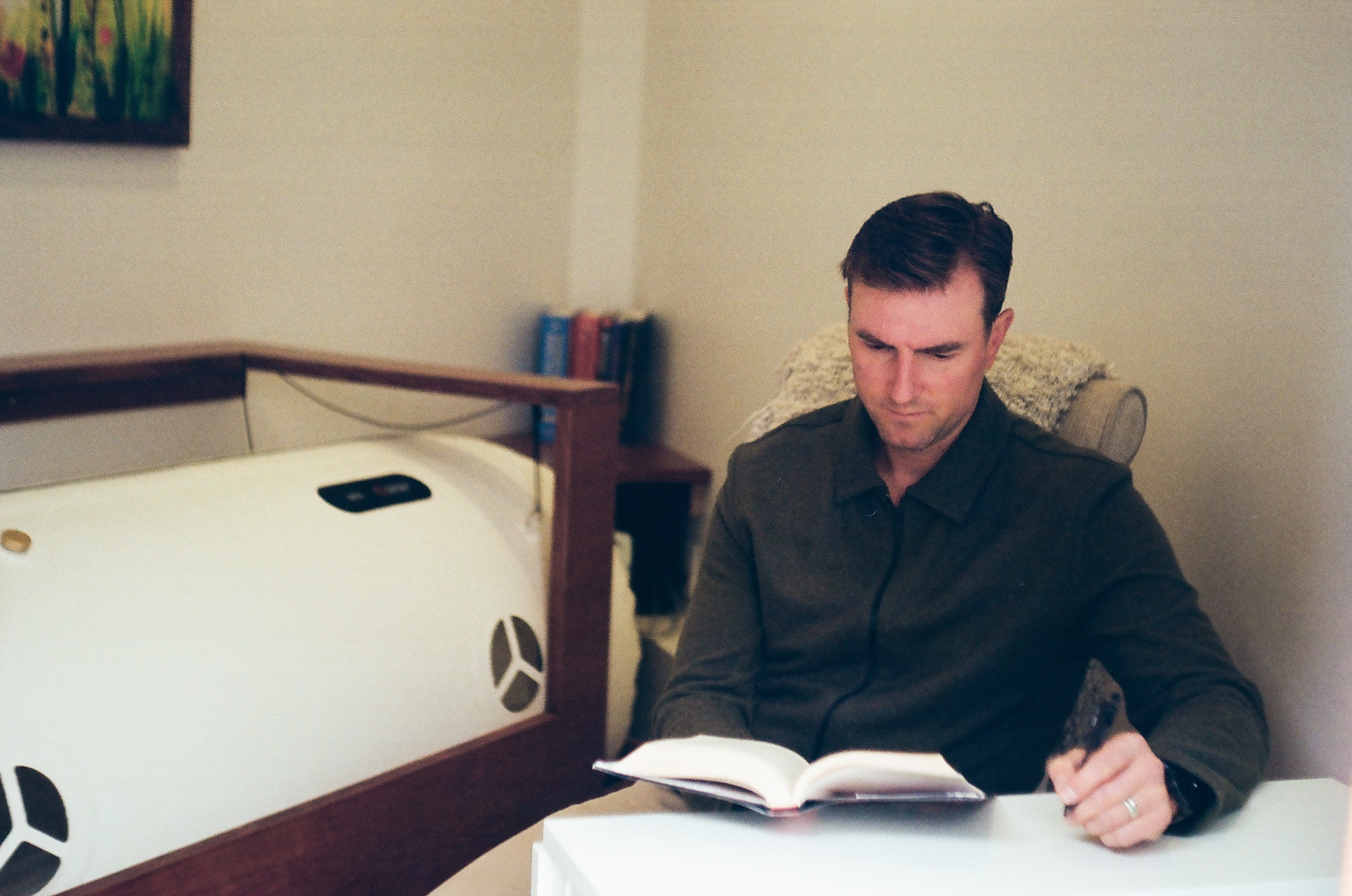 A guest sits quietly with a book beside the soft-shell chamber. Soft natural light, warm wood frame, calm room.